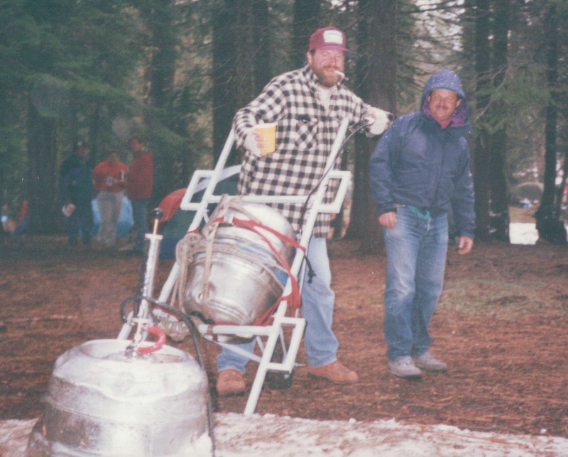 The Wheely stretcher at rest in camp with two kegs — white PVC frame with central rubber wheel, two men standing with it