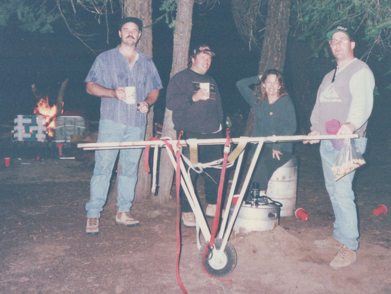 The Wheely stretcher at night — profile view showing the carry frame, wheel, and keg, campfire in background
